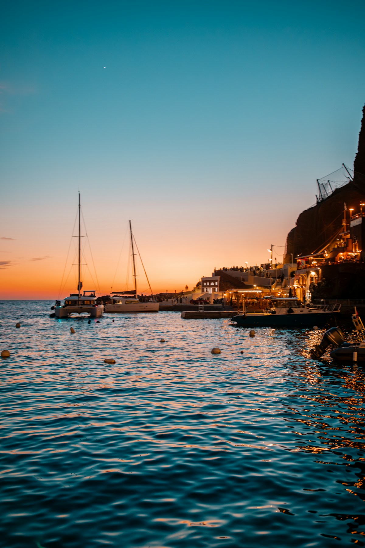 Fira harbour at sunset with sailing boats and illuminated cliff, Santorini, Greece