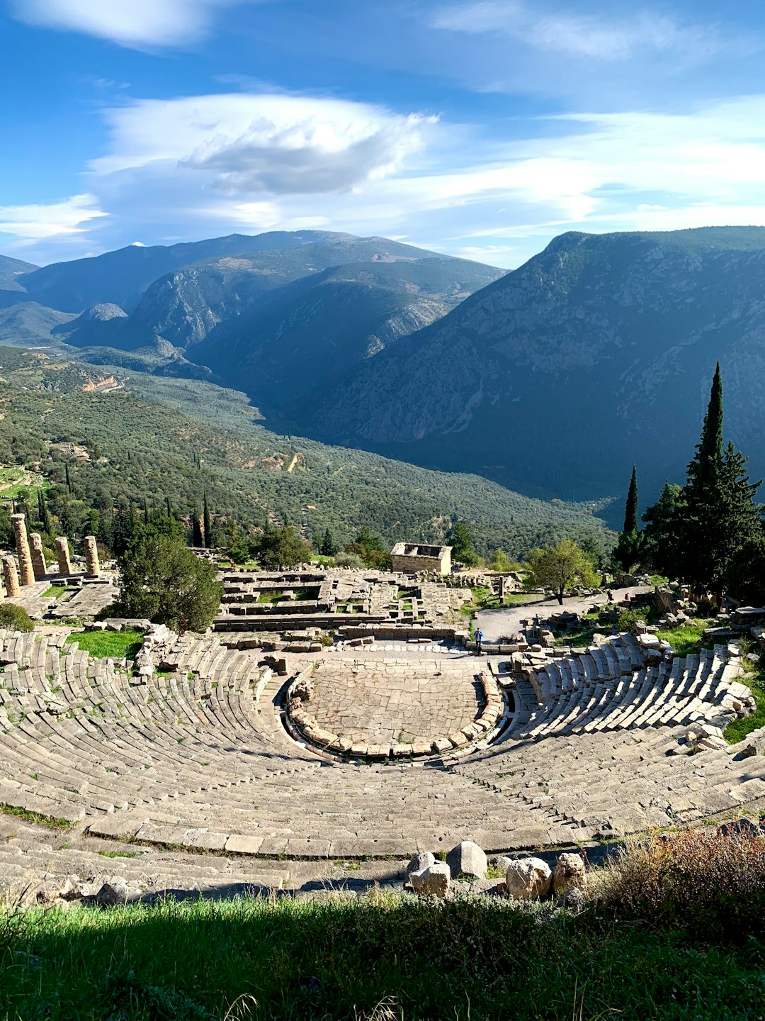 The ancient theatre of Delphi with its stone semicircular seating, columns of the Temple of Apollo below, and the deep green valley of Phocis stretching to the horizon
