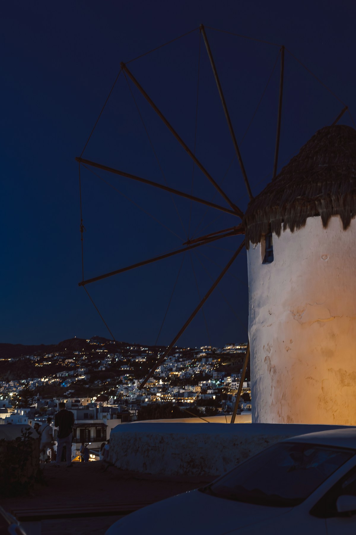 Illuminated white windmill against a deep blue night sky with the lights of Mykonos town glittering below