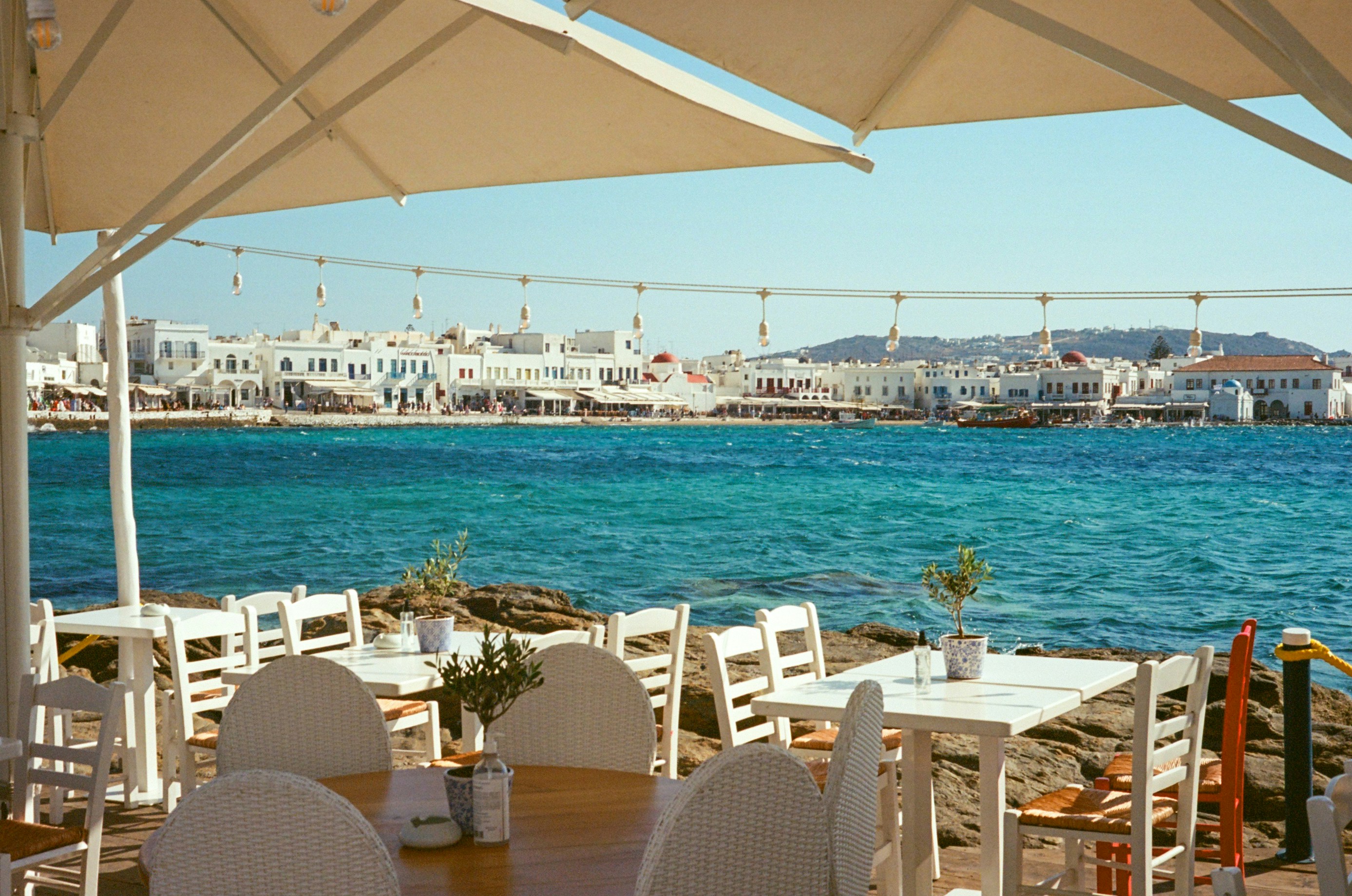 White outdoor dining tables overlooking the turquoise sea and Mykonos town waterfront