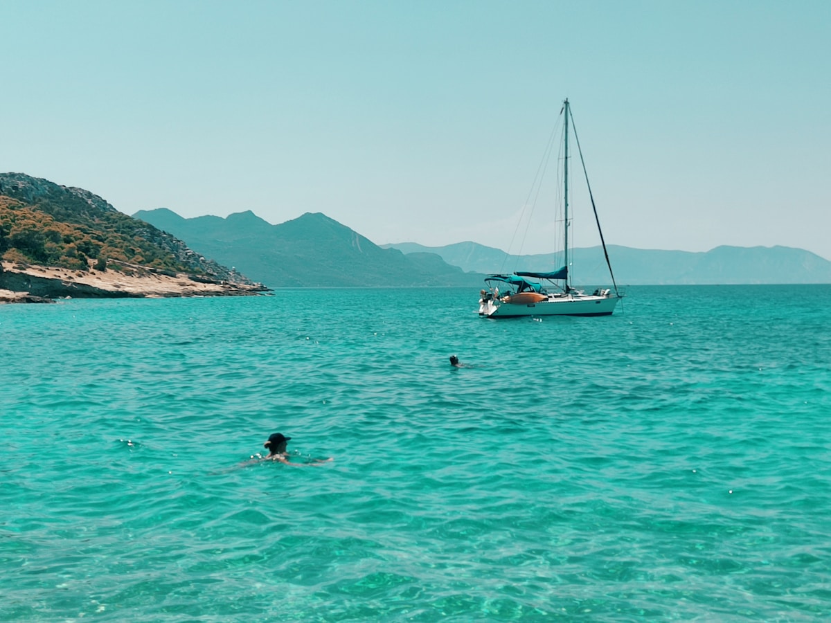 Swimmer in turquoise water with sailboat anchored near green rocky hillside, Aegina, Greece