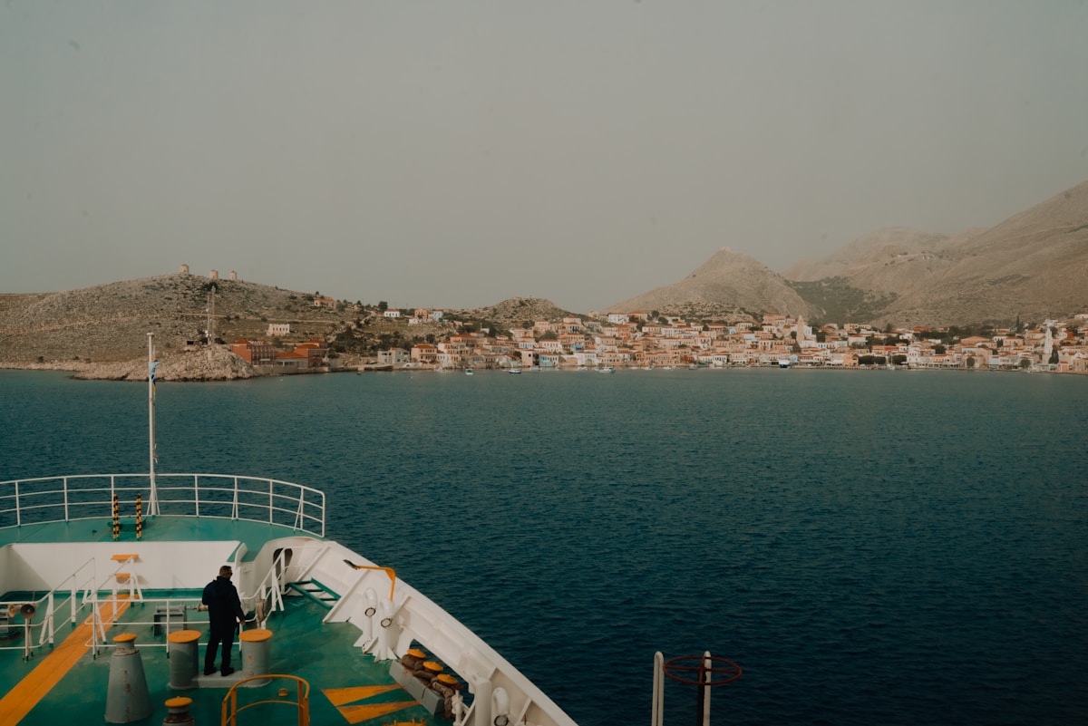 Fähre nähert sich dem farbenfrohen neoklassischen Hafen der Insel Halki, Dodekanes, Griechenland, mit einer Windmühle am Hang