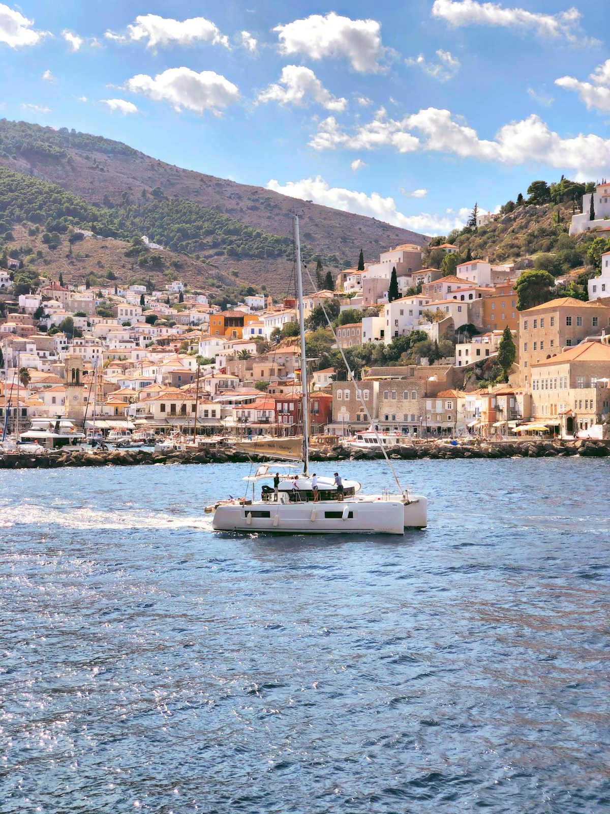 Sailboat approaching Hydra harbour with the town's stone mansions and church clock tower behind, Greece