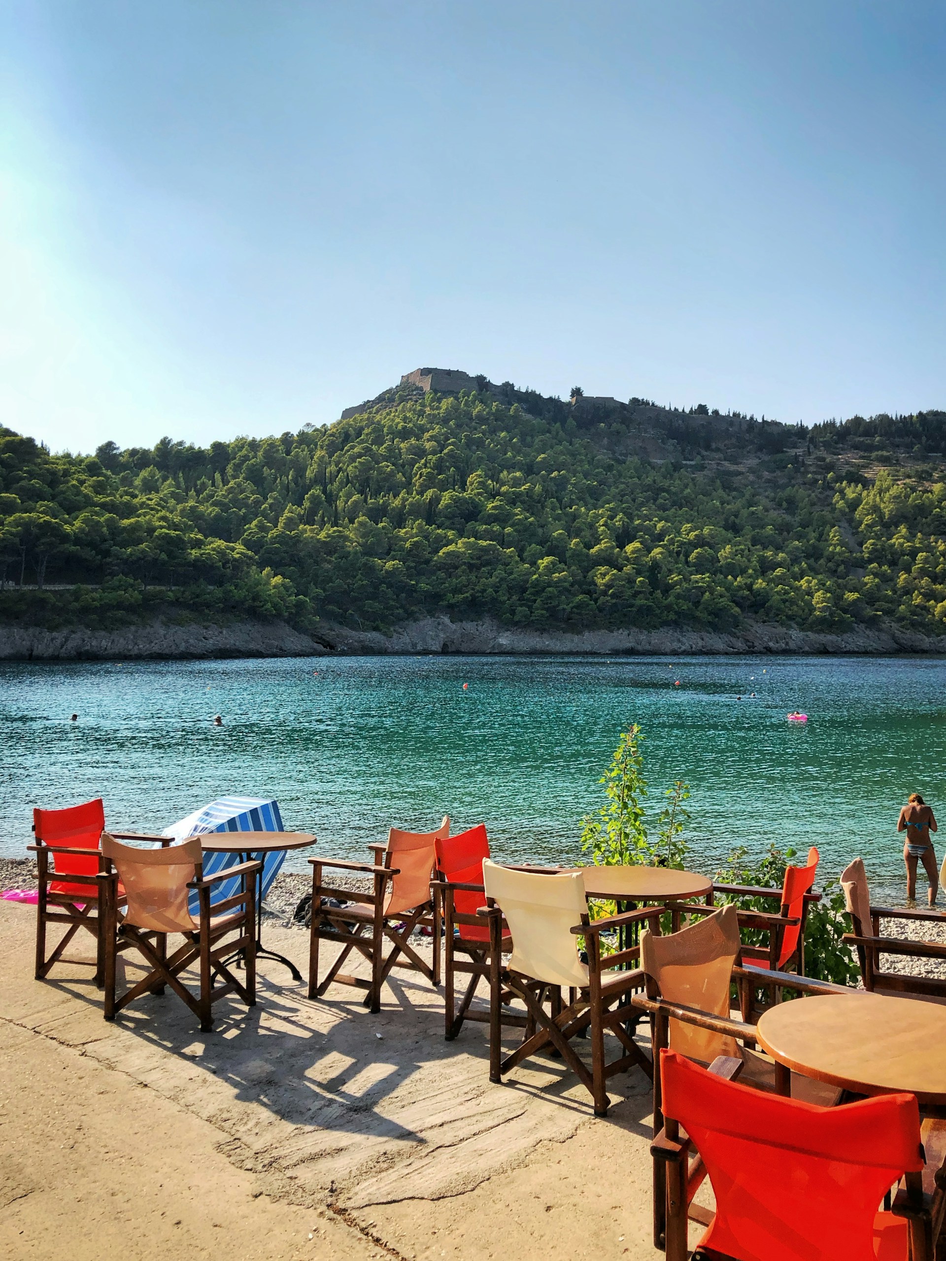 Outdoor dining chairs and tables beside turquoise water with green forested hill behind, Assos, Kefalonia, Greece