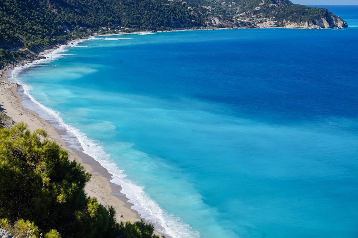 Aerial view of a sweeping turquoise beach in Lefkada, Greece, with pine-covered cliffs and deep blue Ionian water