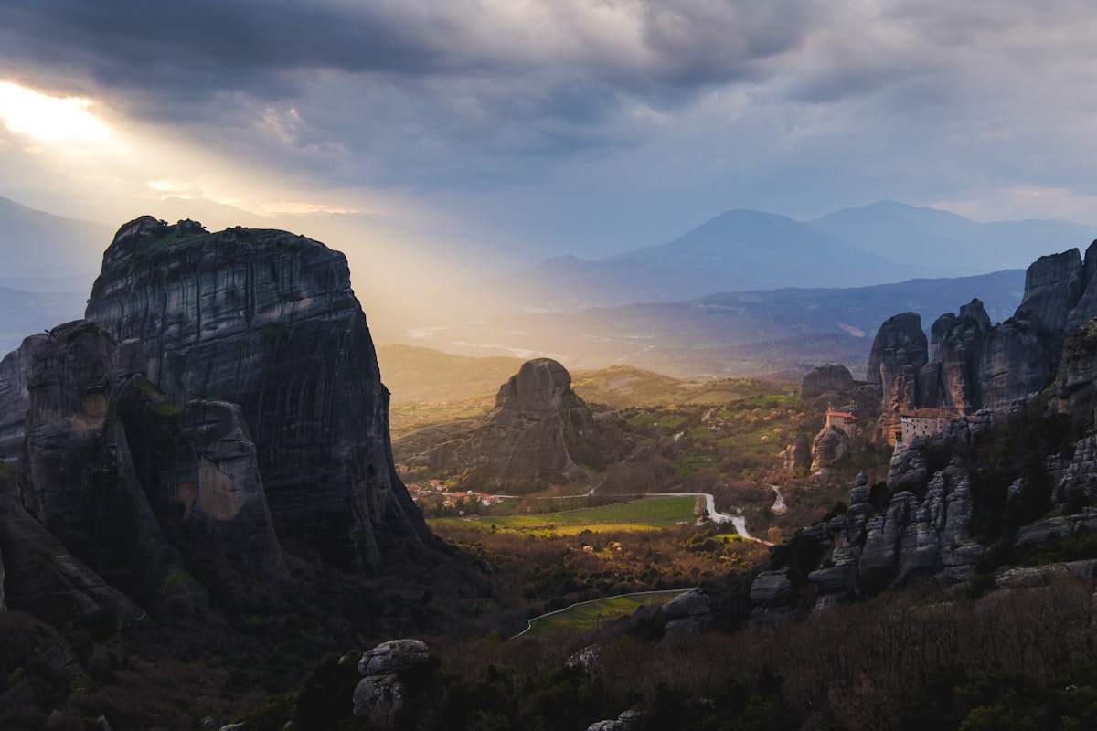 Dramatic rock pillars of Meteora rising above the valley at dusk with monastery visible among the cliffs and mountains beyond