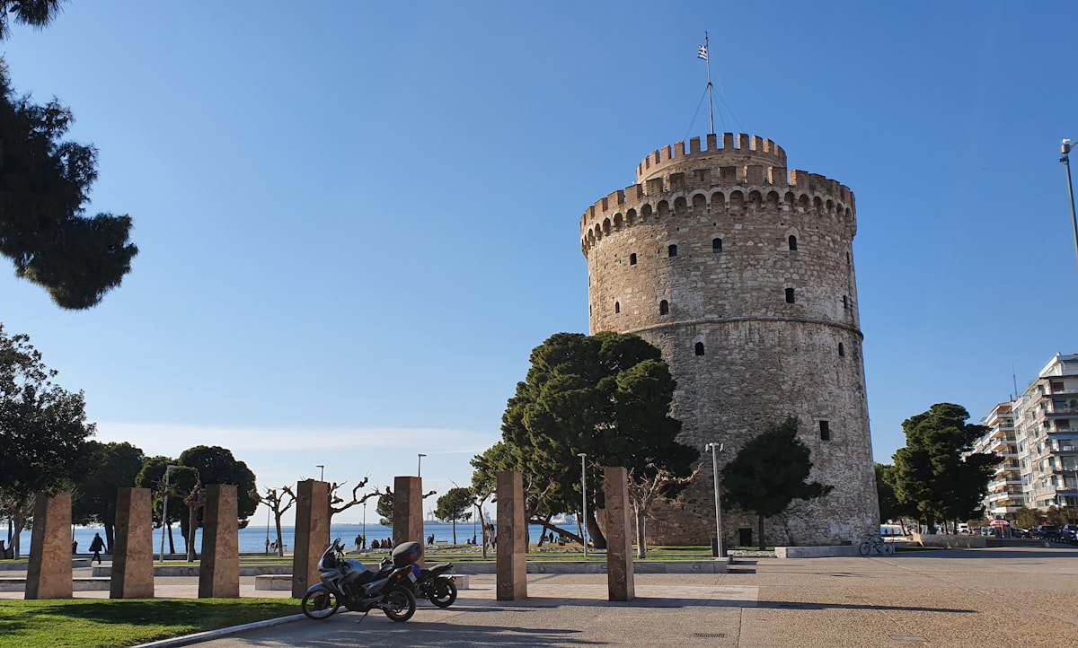 The White Tower of Thessaloniki on the waterfront promenade with the Greek flag and Thermaic Gulf behind it